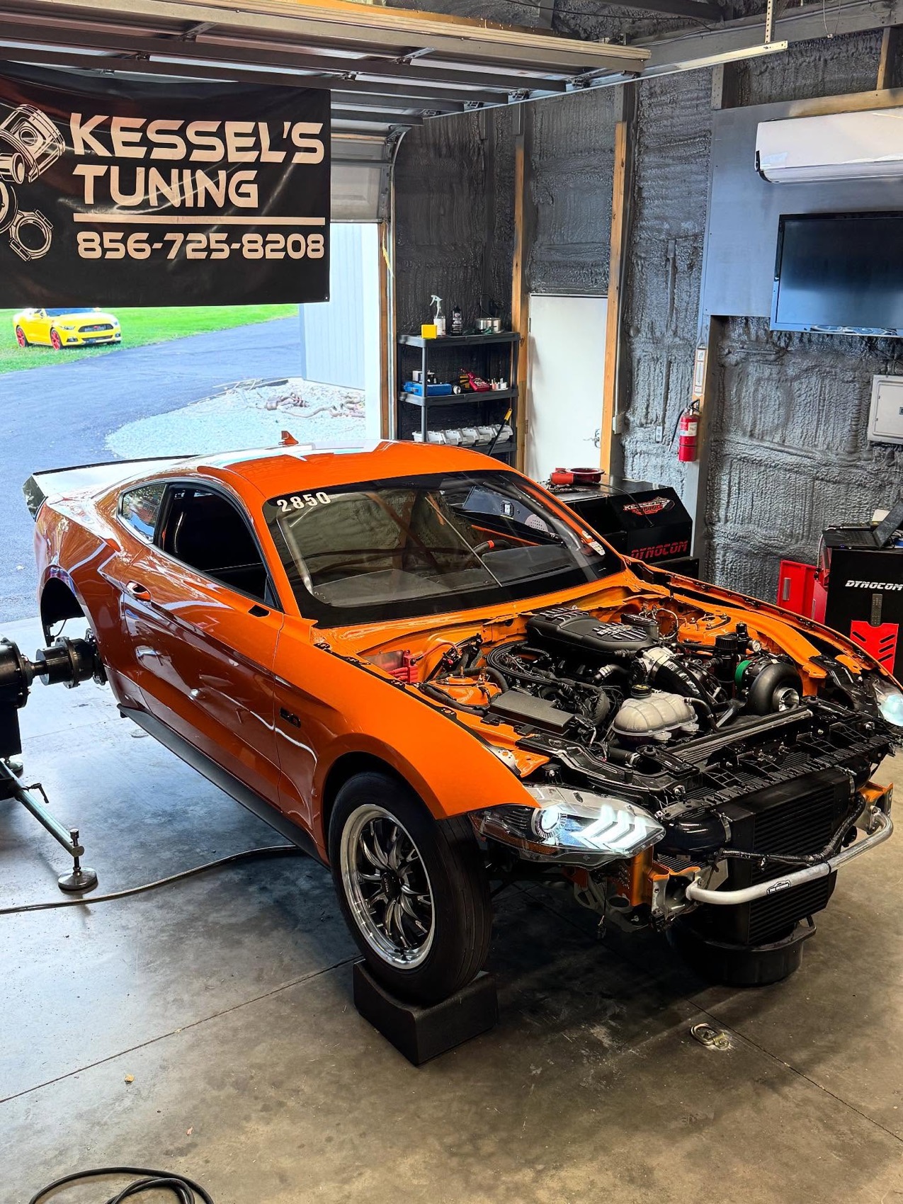 An orange Mustang with the front end stripped and a turbo fitted, sitting on the hub dyno under the Kessel's Tuning banner