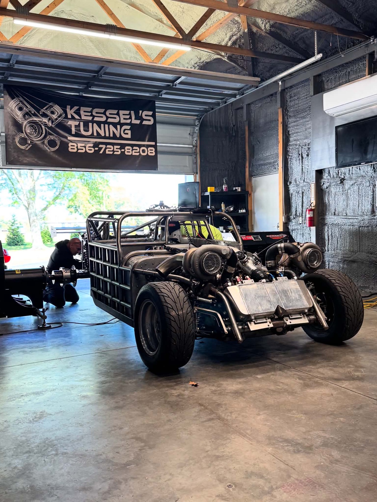 A bare metal twin turbo tube chassis car in the shop bay under the Kessel's Tuning banner