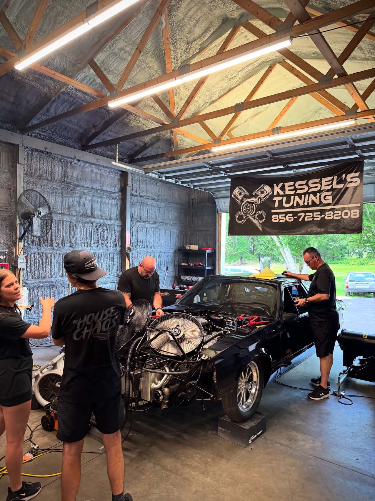Four people around a black GM coupe with the front end open on the dyno, under the Kessel's Tuning banner
